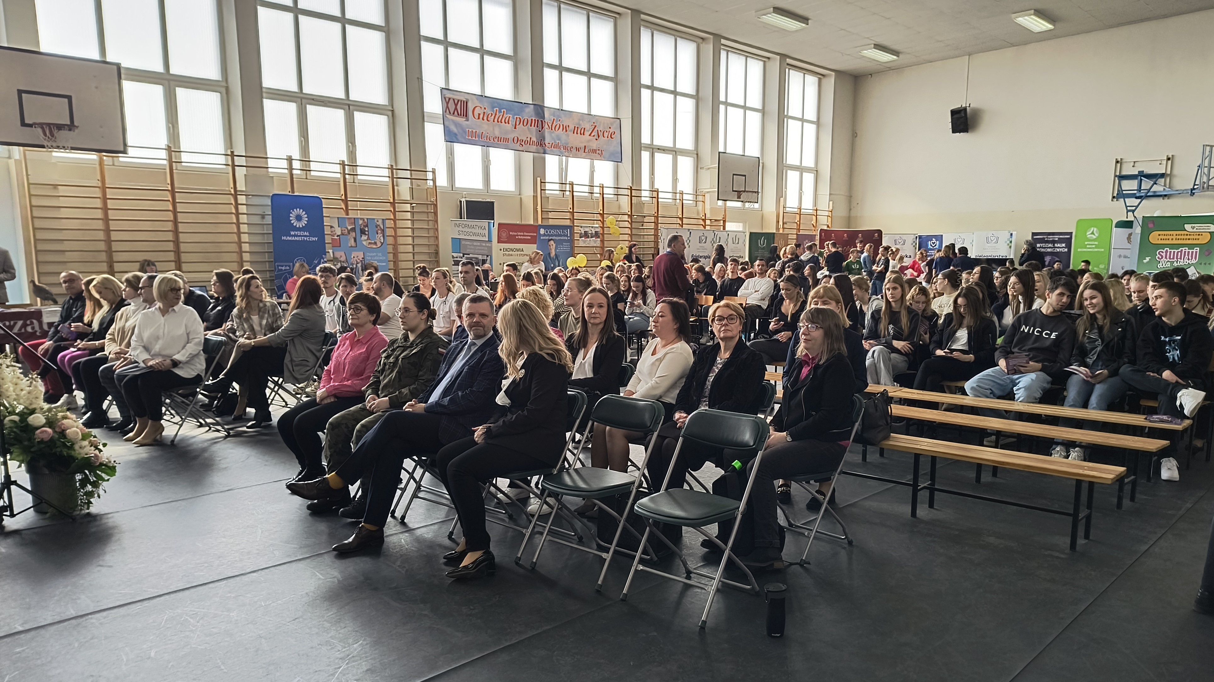 Ludzie siedzący na krzesłach w szkolnej sali gimnastycznej.