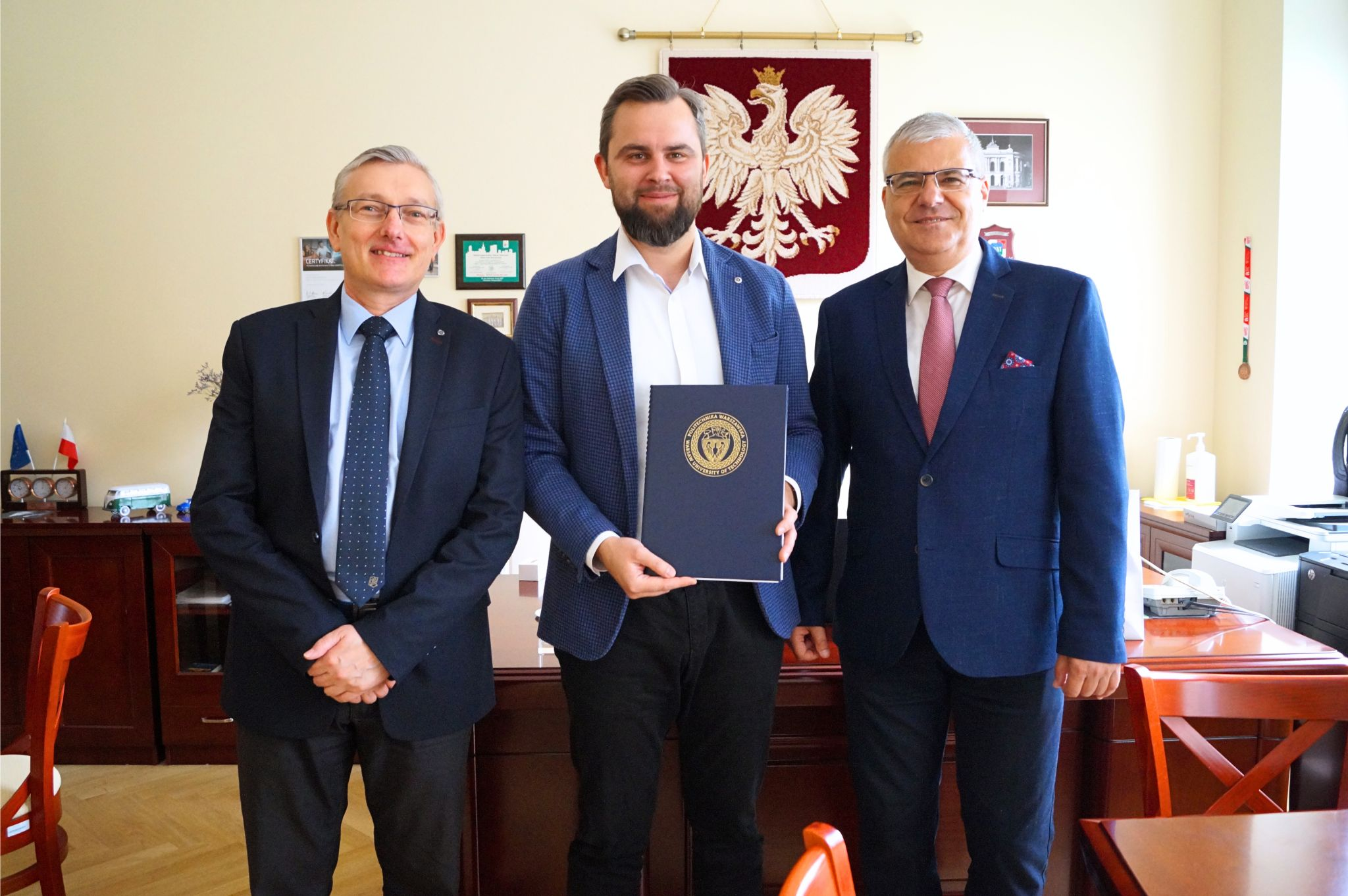 Fotografia trzech mężczyzn na tle biurka i godła Polski na ścianie